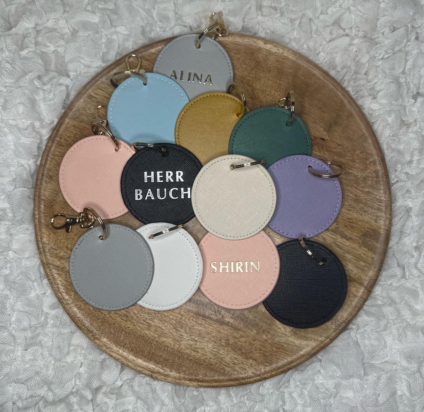 Auf dem weißen Hintergrund befindet sich ein Holztablett. Auf dem Tablett liegen Schlüsselanhänger in verschiedenen Farben & ein paar mit in Gold & Silber geprägte Namen darauf.