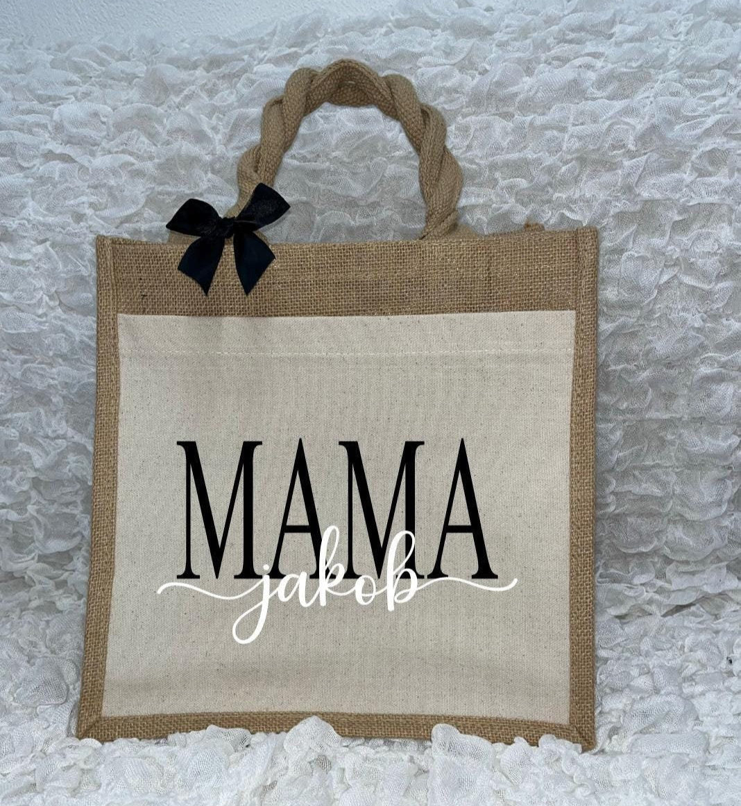 Vor dem weißen Hintergrund befindet sich ein Juteshopper mit einer fast vollflächigen Baumwolltasche darauf. Links auf dem Henkel befindet sich eine schwarze Schleife. Der personalisierte Aufdruck besteht aus Mama & dem/den Wunschnamen.