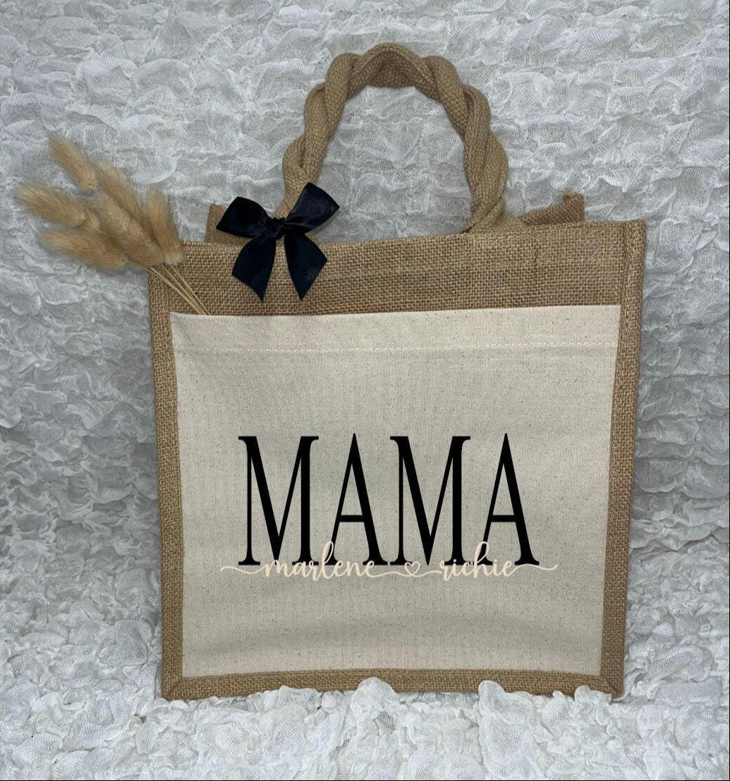 Vor dem weißen Hintergrund befindet sich ein Juteshopper mit einer fast vollflächigen Baumwolltasche darauf. Links auf dem Henkel befindet sich eine schwarze Schleife. Der personalisierte Aufdruck besteht aus Mama & dem/den Wunschnamen.