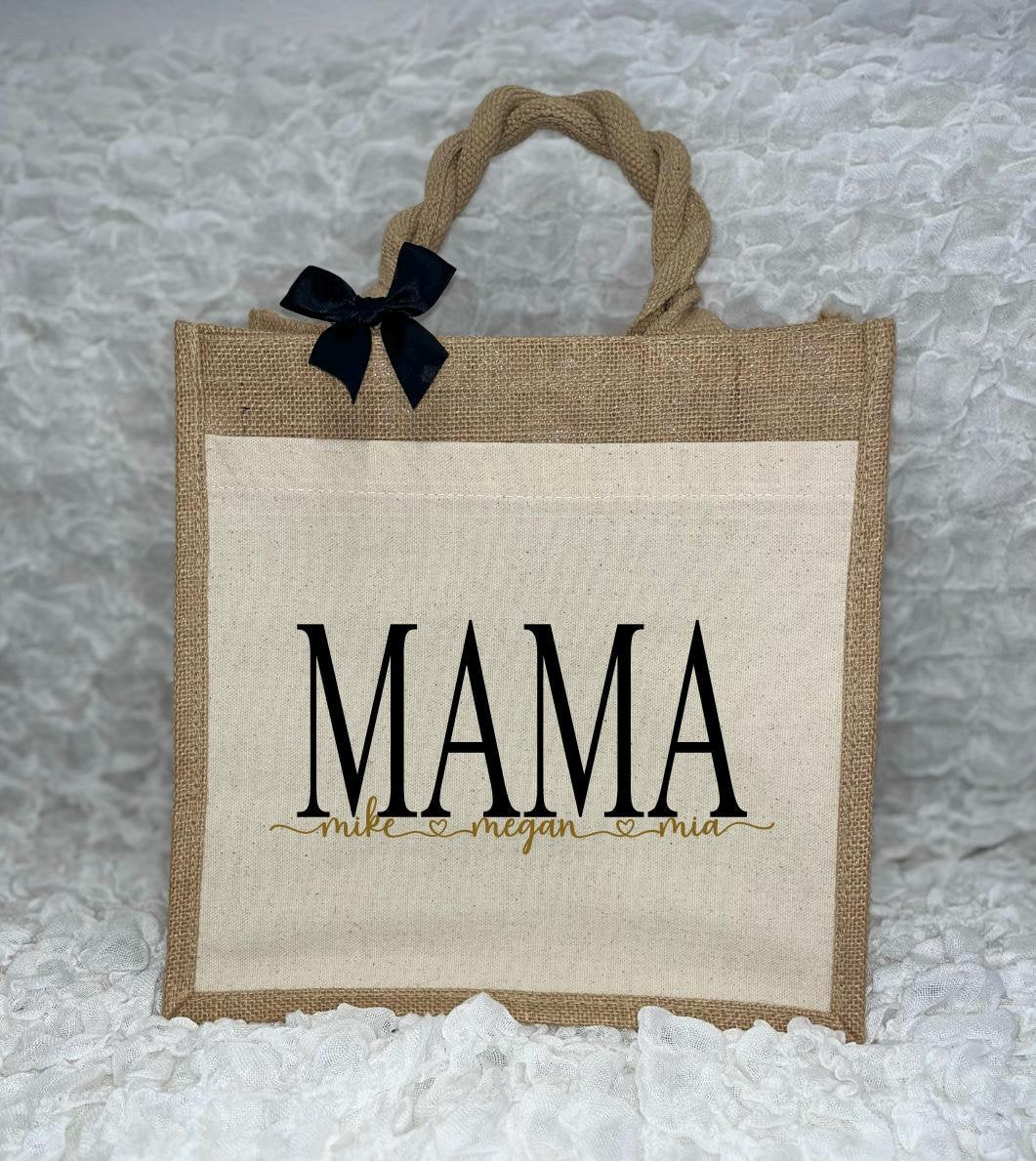 Vor dem weißen Hintergrund befindet sich ein Juteshopper mit einer fast vollflächigen Baumwolltasche darauf. Links auf dem Henkel befindet sich eine schwarze Schleife. Der personalisierte Aufdruck besteht aus Mama & dem/den Wunschnamen.